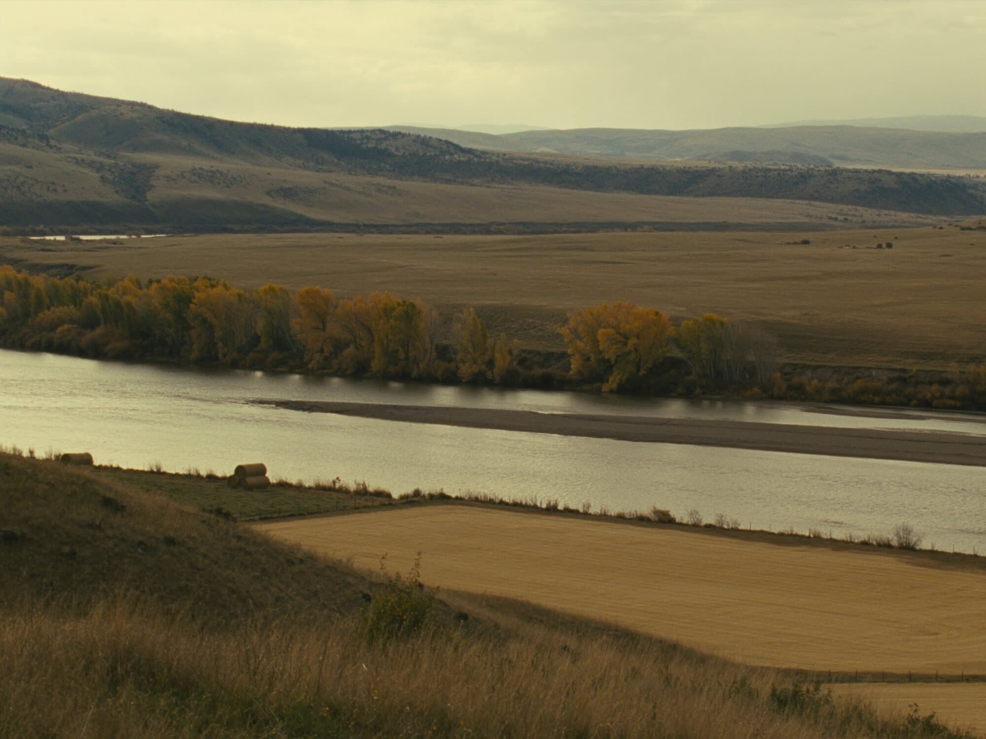 A wide river bend lined with cottonwoods turning gold, ranch hayfields and dry foothills beyond.