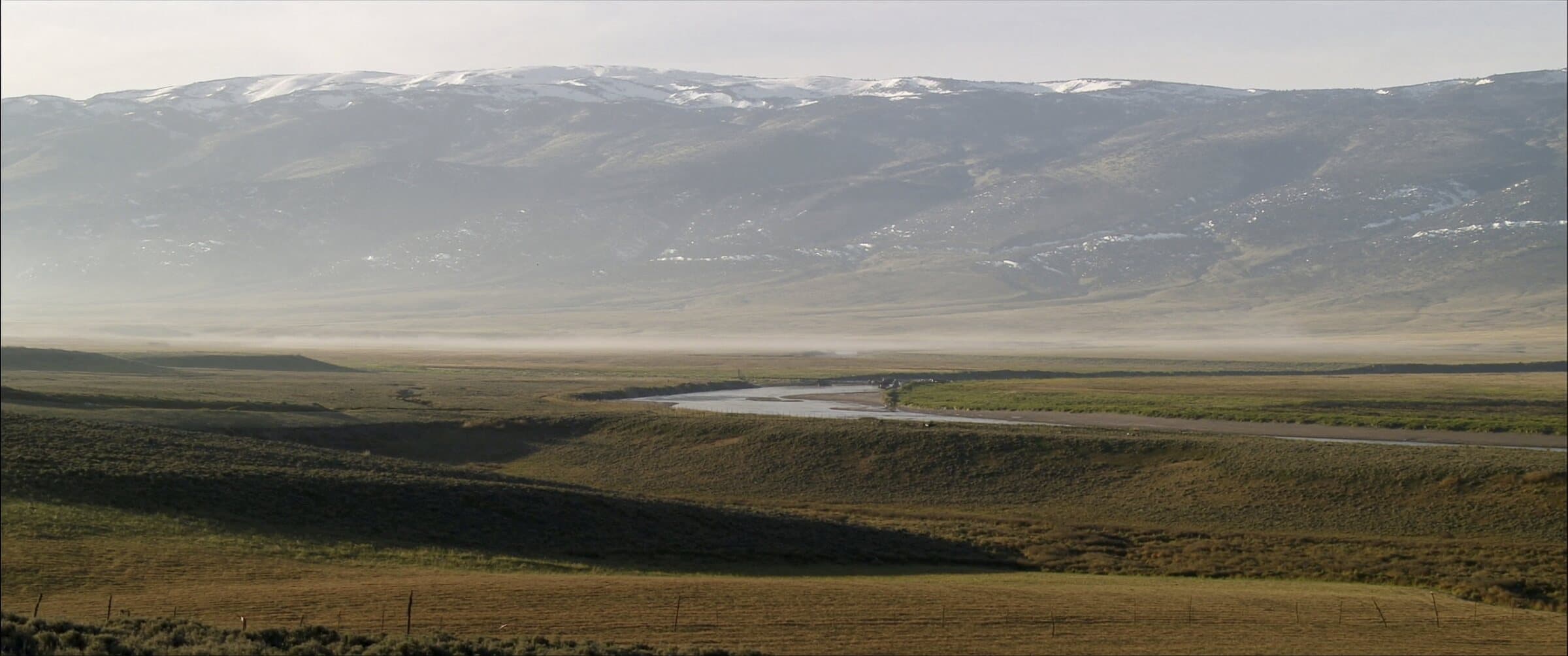 A wide Western ranch valley at first light, a river glinting through hayfields below snow-dusted peaks.