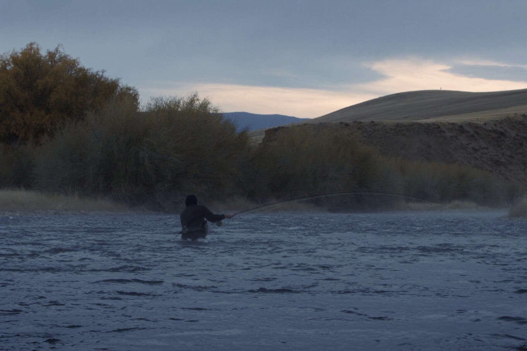 A fly angler wading a wide Western river at first light, steam lifting off the water.